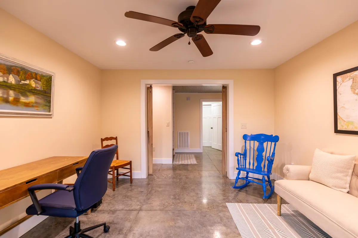 A cozy sitting room features a blue rocking chair by a wooden desk, a cushioned sofa, ceiling fan, and art on the wall. The setting is comfortable and conducive to reading, relaxing, or productive work, with peaceful views over the grounds.