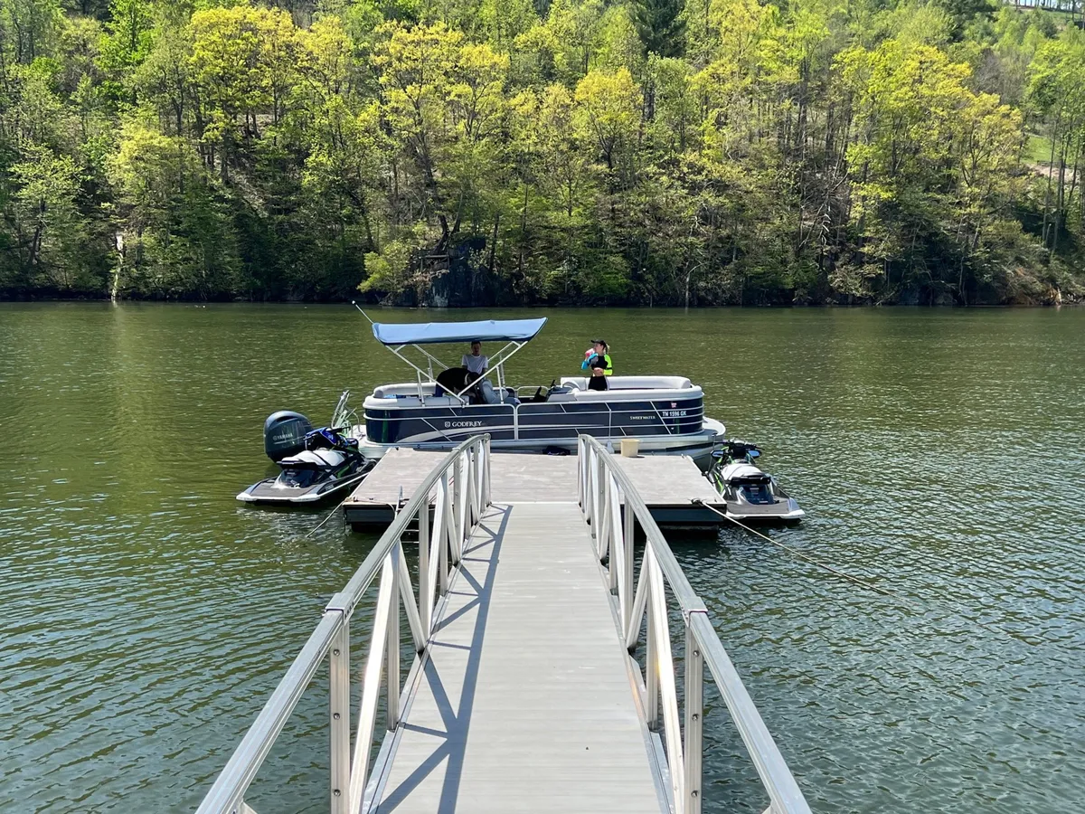 A serene lakeside scene featuring a dock with a boat moored, calm water, and lush trees surrounding the area. The atmosphere is relaxed and inviting, with the dock providing direct access to boating and water activities.