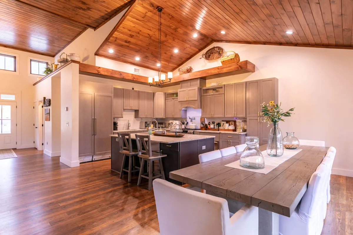 This cozy, open-plan space seamlessly showcases a kitchen island, a large dining table, and rich wooden ceiling beams. The dining area invites group meals and lively conversation, while the kitchen remains visually connected to the rest of the living space.
