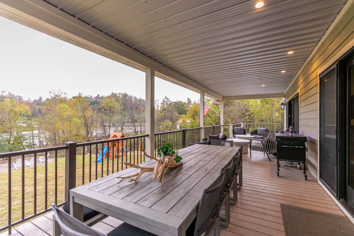 A large outdoor deck with a spacious dining table and comfortable seating, set against a scenic river view. The background features a playground, blending adult relaxation with family-friendly fun.