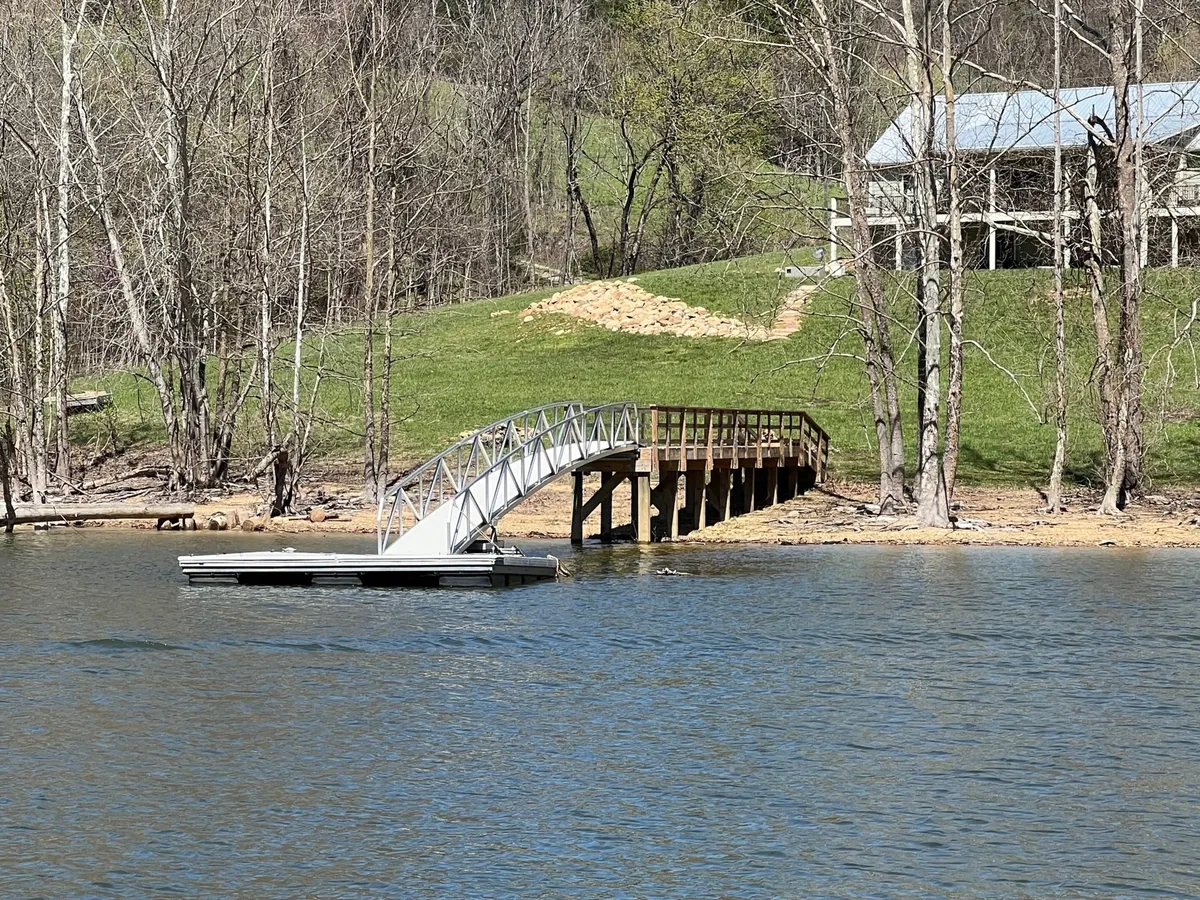 A tranquil exterior setting with a wooden bridge leading to a floating dock extending over the water. The gentle slope to the dock and calm surface invite fishing, boating, or a peaceful picnic, with a beautiful view across the lake.