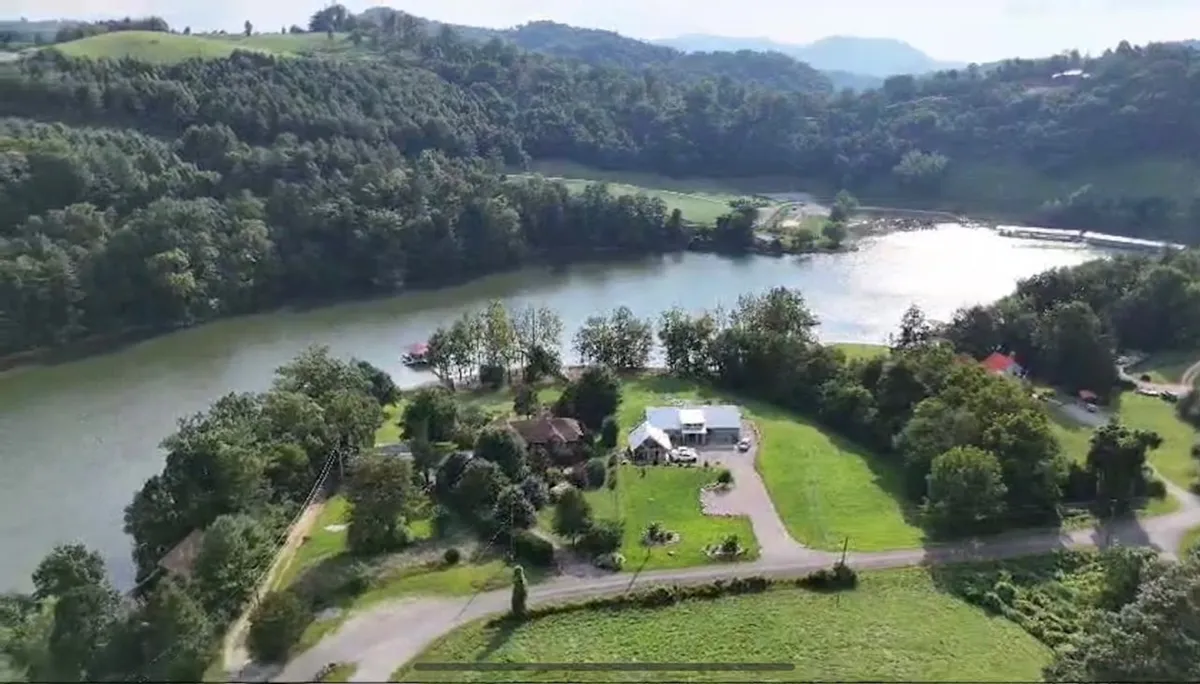 The hero image showcases a luxurious lakeside home set against the tranquil waters of Watauga Lake, with lush green fields and rolling hills in the background. The scene captures a peaceful sunset reflected on the water, hinting at evenings by the lake, and an inviting landscape perfect for outdoor activities like fishing, boating, and relaxing in nature.