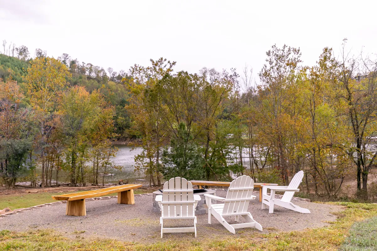A serene setup of Adirondack chairs circled around a fire pit with a river view and natural surroundings. The image suggests tranquil evenings spent outdoors, socializing or simply soaking in the beauty of the landscape.
