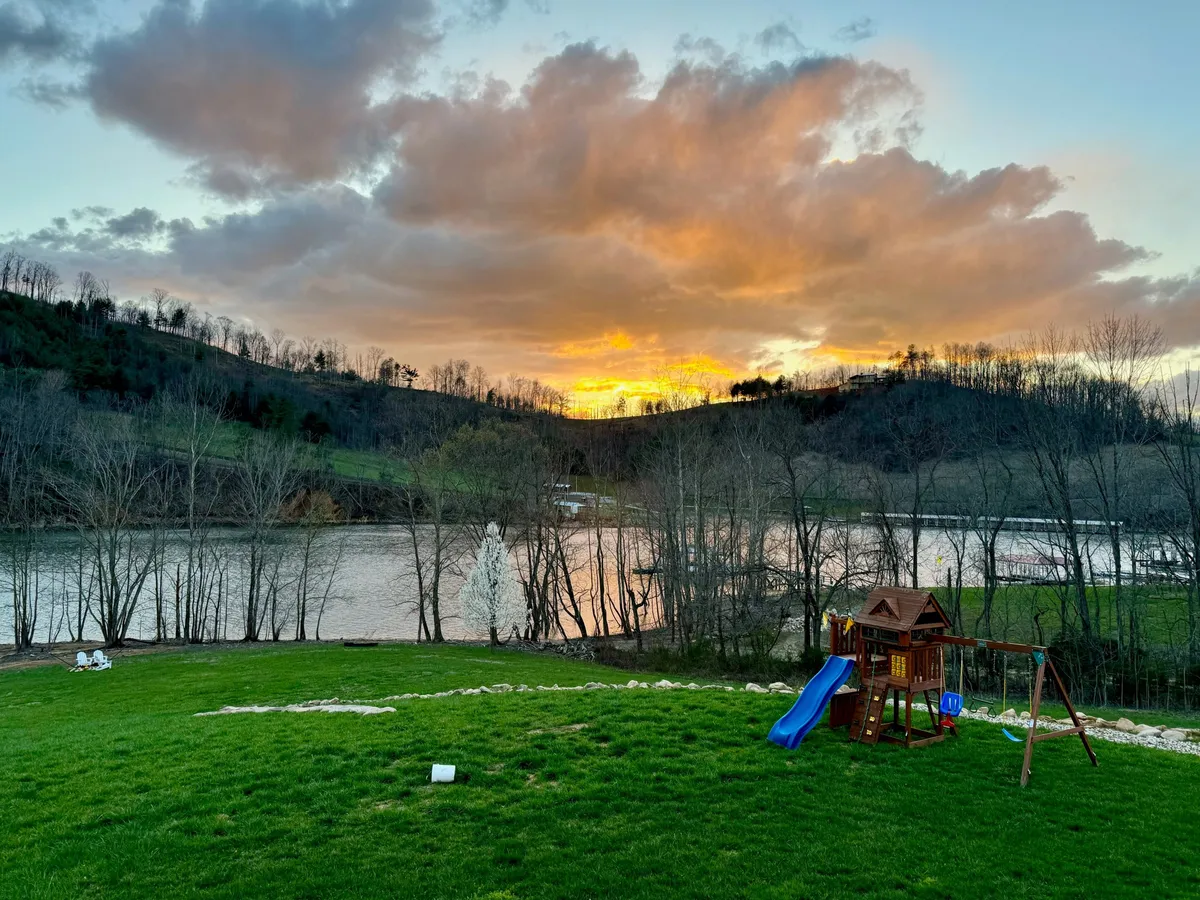 An inviting outdoor scene featuring a playground set, a flowing river, rolling hills, and a glowing sunset. The expansive lawn creates opportunities for group activities and family fun, while the water and horizon promise tranquil evenings and relaxation.