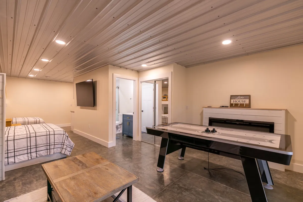 A multifaceted retreat space highlighted by an air hockey table, a bed, a TV, and a fireplace, with a cozy coffee table at the center. The arrangement suggests both entertainment and comfort, with the air hockey table ready for after-dark tournaments and the bed and fireplace offering options for winding down.