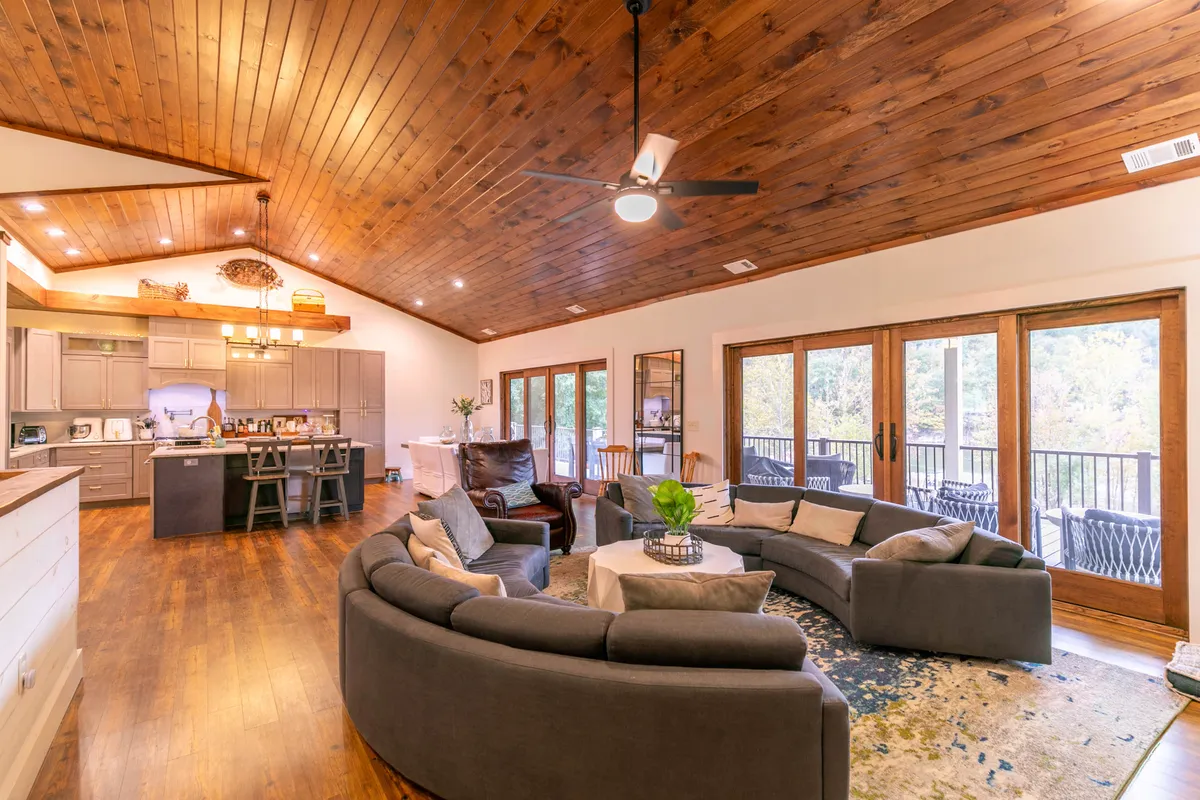 A sunlit, open-concept living space featuring a curved sectional sofa, large windows with tranquil outdoor views, visible kitchen island, and wooden beam ceilings. The room exudes warmth, comfort, and a sense of airiness, perfect for family gatherings or relaxing by the lake.
