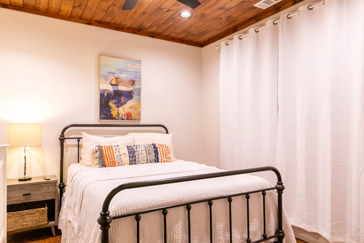A beautifully made bed is the centerpiece of this inviting bedroom, complemented by elegant artwork, a cozy lamp, and soft curtains that filter gentle natural light. The overall feel is one of tranquility and comfort, perfect for restful nights or reading in bed.